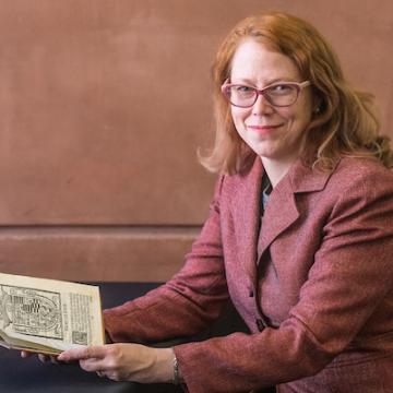 a white woman with red medium length hair and glasses, wearing a rose colored blazer sits at a table holding a book