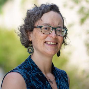 a white woman with short wavy graying brown hair and glasses is smiling in front of an out of focus outdoor background