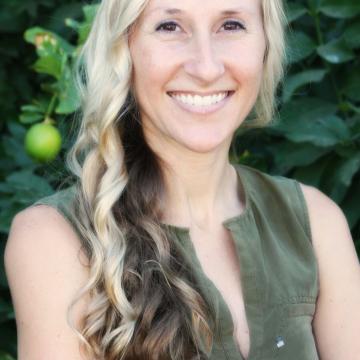 a light-skinned woman with long blond and brown hair in a green sleevless top stands with arms crossed in front of a citrus tree